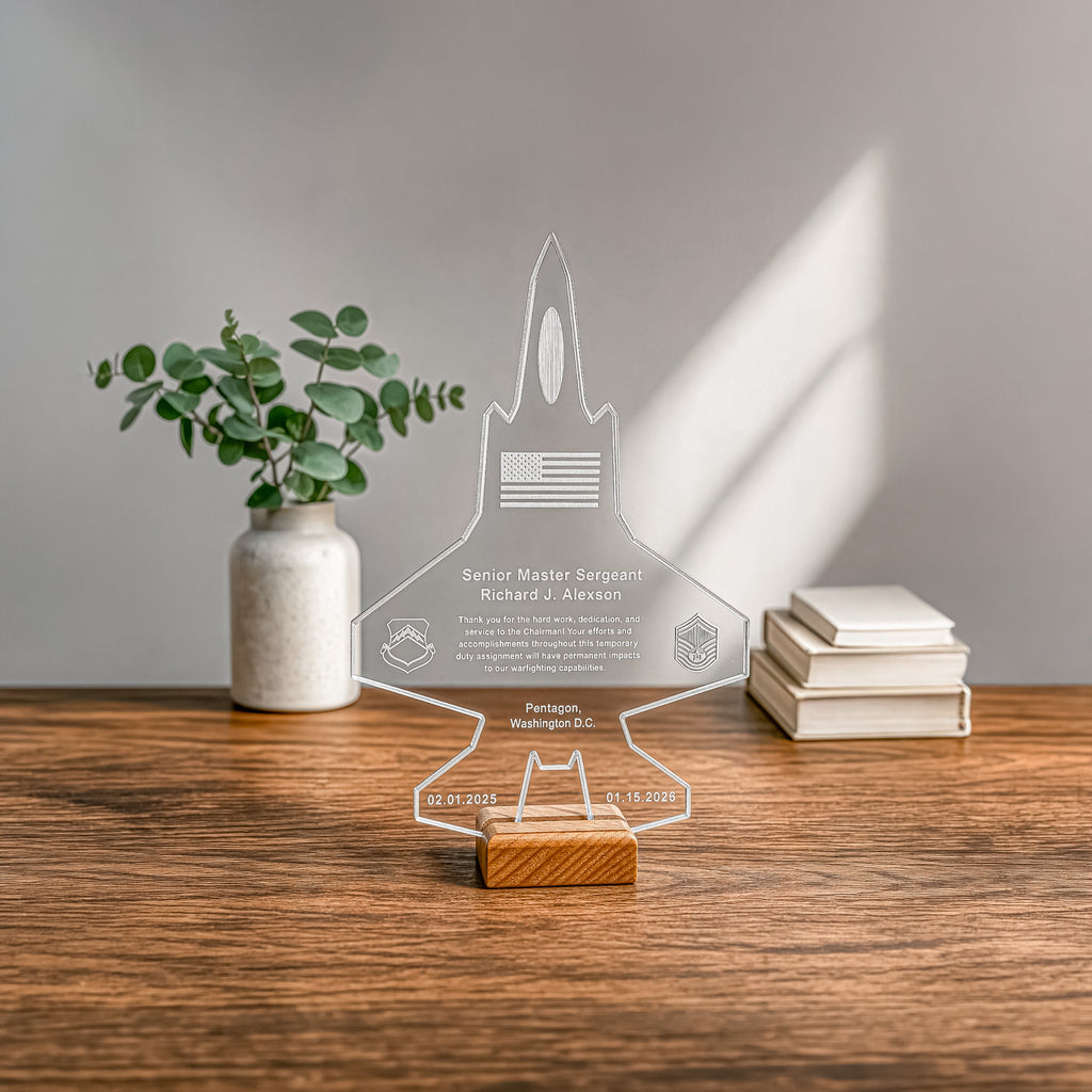 A clear acrylic military-style jet-shaped keepsake plaque with engraved text and insignia, displayed on a small wooden block on a wooden tabletop. A white ceramic vase with green stems and a stack of books sit blurred in the background against a light gray wall with soft window-light shadows.