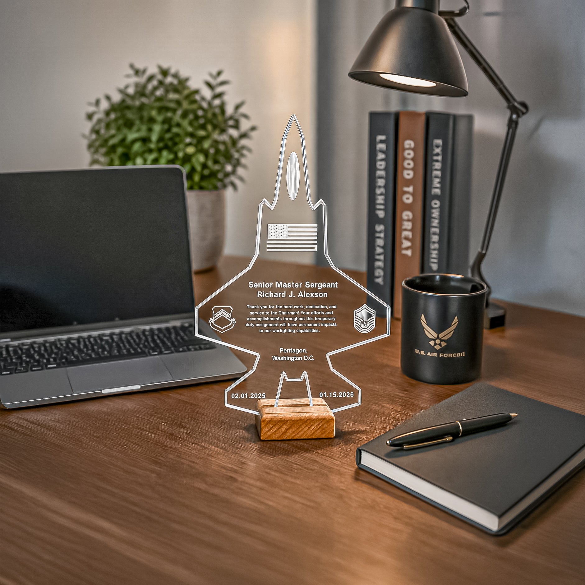 A clear acrylic personalized rocket-shaped display stand on a wooden base, placed on a desk with a laptop, lamp, books, mug, notebook, and pen in a real indoor setting.