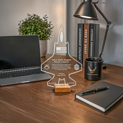 A clear acrylic personalized rocket-shaped display stand on a wooden base, placed on a desk with a laptop, lamp, books, mug, notebook, and pen in a real indoor setting.