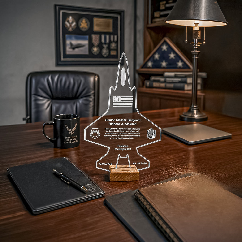 A personalized acrylic military achievement plaque shaped like a fighter jet, displayed on a wooden desk in a professional office setting alongside a black leather chair, a black mug with the U.S.