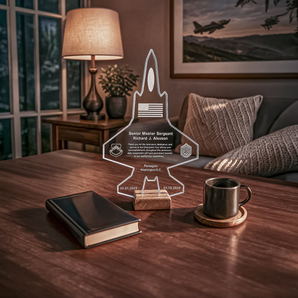 A clear acrylic personalized plaque shaped like a fighter jet stands on a wooden block on a dark wood table in a cozy living room setting. The plaque features custom engraved text and insignia.