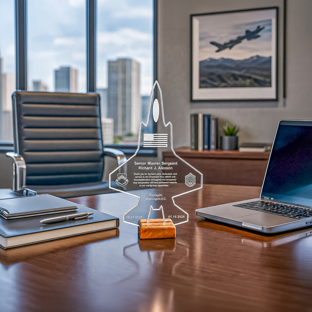A clear acrylic custom-engraved trophy plaque shaped like a fighter jet stands on a small orange base on a polished wooden desk in a modern office setting. The background features a black leather office chair, a laptop, a notebook, and a framed picture of a military jet on the wall.