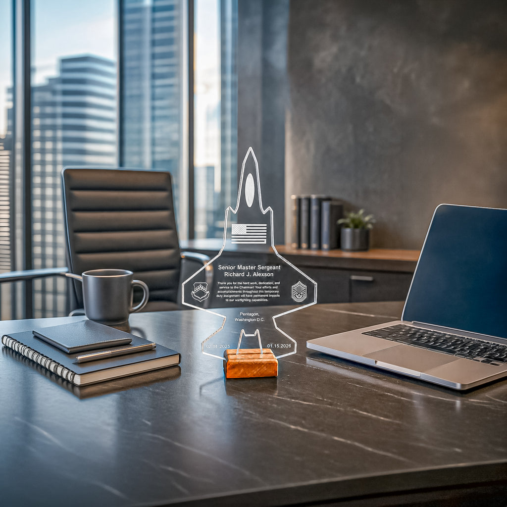 A clear acrylic personalized rocket-shaped award plaque stands on a small orange block on a dark office desk, with a laptop, notebook, and coffee mug nearby in a modern office setting.