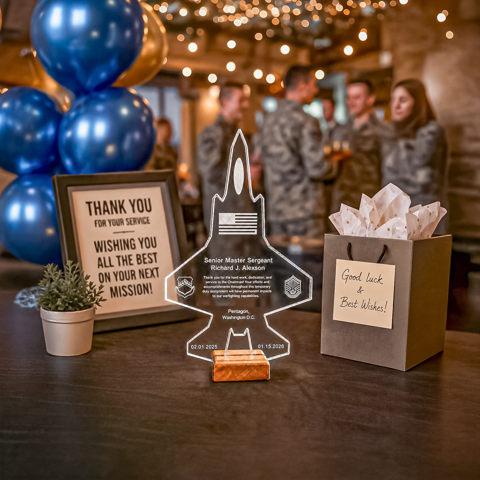 A personalized acrylic jet-shaped award plaque stands on a dark wooden table in a party setting, flanked by a framed thank-you sign, a small potted plant, and a gift bag with a 'Good luck & Best Wishes!' note. The background shows blurred people and blue balloons.