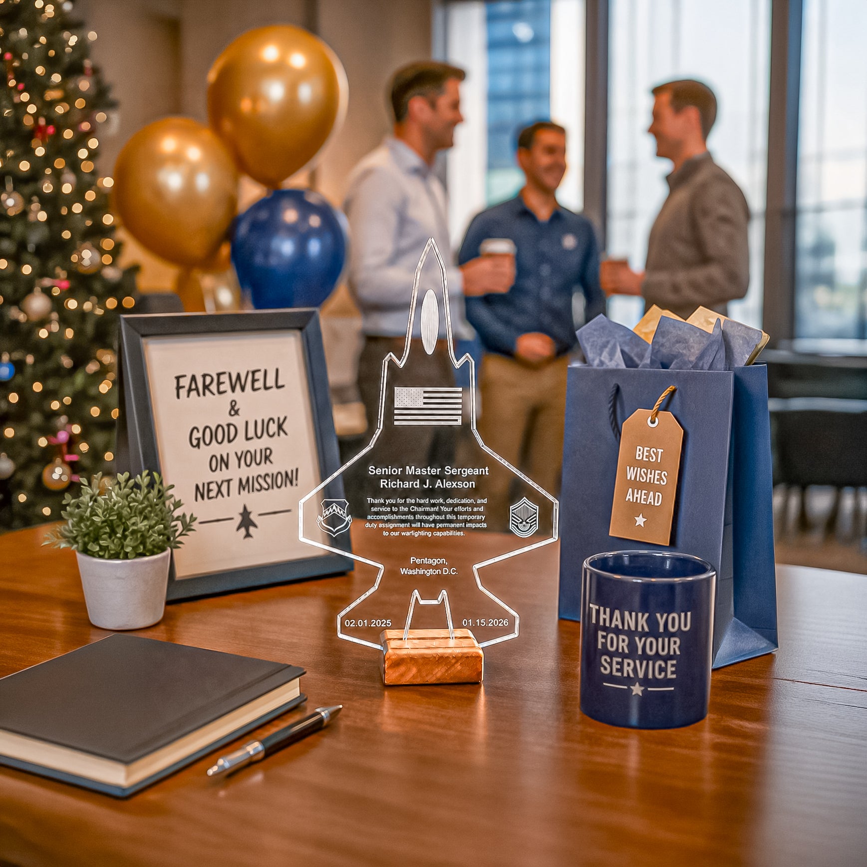 A styled farewell gift set on a wooden table, featuring a clear acrylic jet-shaped keepsake plaque, a navy blue mug reading 'THANK YOU FOR YOUR SERVICE', a navy gift bag with a 'BEST WISHES AHEAD' tag, a framed sign that says 'FAREWELL & GOOD LUCK ON YOUR NEXT MISSION!', a notebook, a pen, and a small potted plant. The background shows a blurred office environment with three men in suits, balloons, and a Christmas tree.
