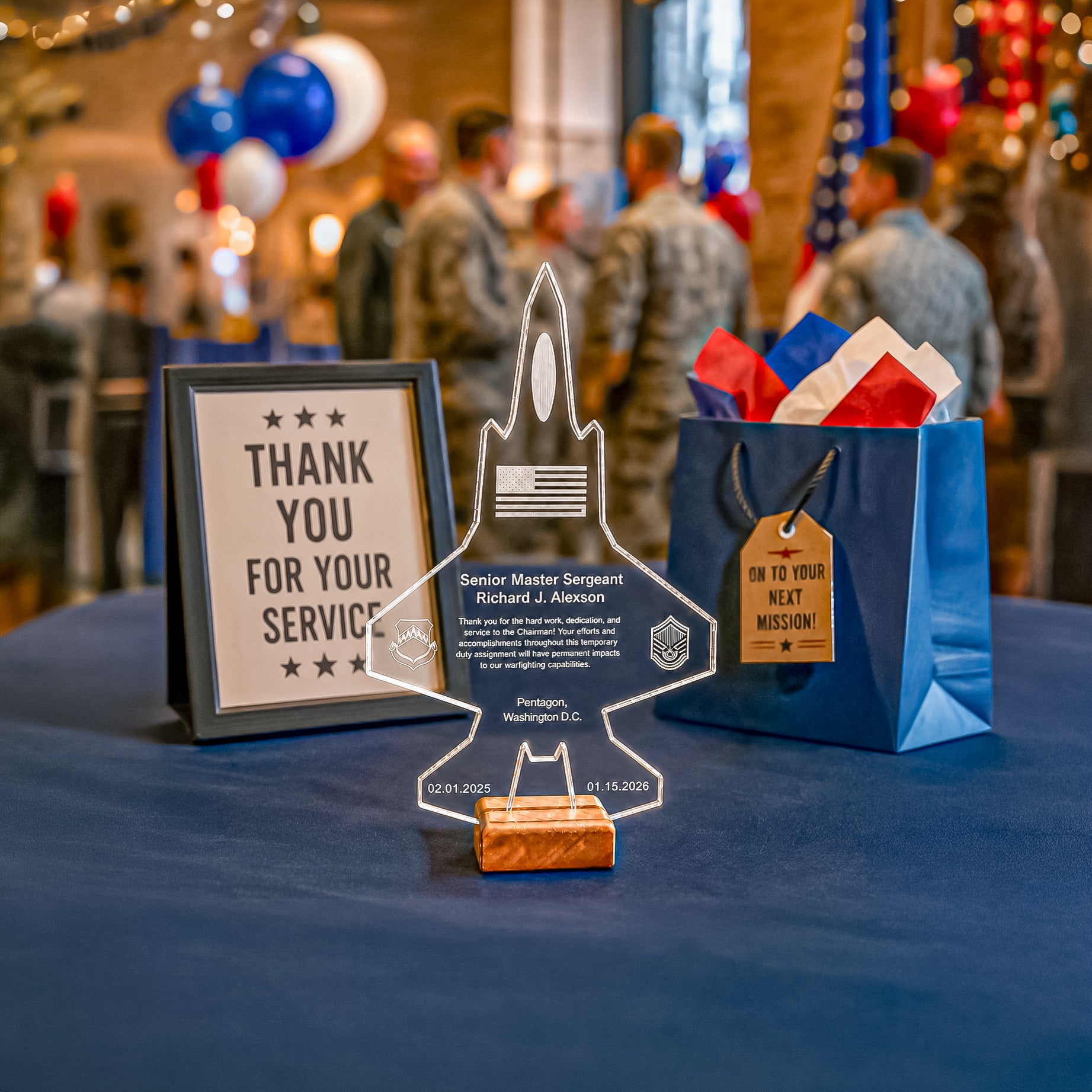 A military commendation acrylic plaque shaped like a fighter jet stands on a table at a ceremony, next to a framed 'Thank You for Your Service' sign and a blue gift bag with a 'On to Your Next Mission' tag. The background shows blurred attendees in military uniforms and balloons.