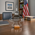 A clear acrylic jet-shaped keepsake plaque with engraved personal details and a small wooden base, displayed on a wooden desk in an office setting with a U.S.