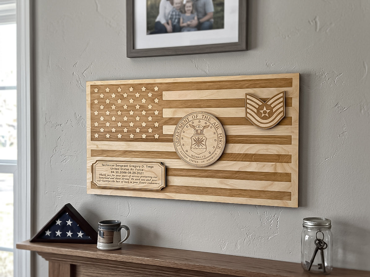 A wooden military memorial plaque featuring a stylized American flag design with engraved stars, stripes, a circular 'Department of the Army' seal, and a rank insignia. The plaque is mounted on a textured gray wall above a wooden mantel with a framed photo, a folded flag, a mug, and a glass jar.
