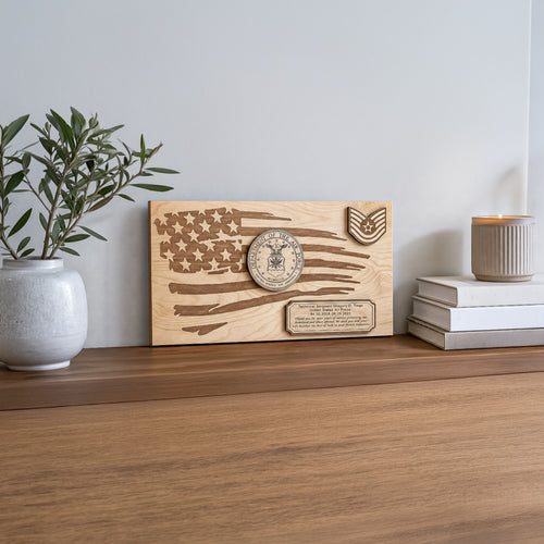 A rectangular wooden plaque featuring an engraved distressed American flag design, military-style insignia, and a small plaque at the bottom, displayed on a wooden surface in a styled interior setting with a plant and books.